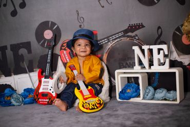 Smiling toddler in a blue hat holding a toy guitar in a music-themed setup.