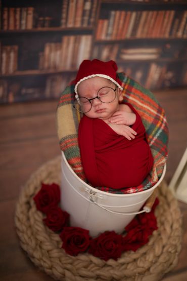 Newborn baby wrapped in red, wearing glasses, sleeping in a white bucket surrounded by red roses.