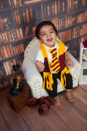 Smiling toddler dressed in a yellow and maroon scarf and tie, sitting on a white armchair.