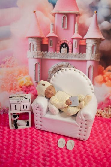 A newborn baby sleeping on a white mini sofa with a pink castle backdrop.