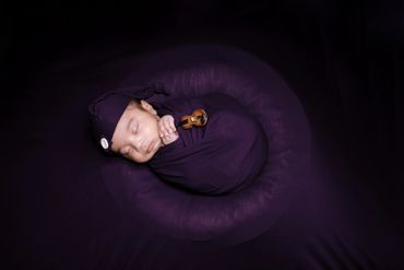 Newborn baby wrapped in purple cloth, holding a tiny violin prop.
