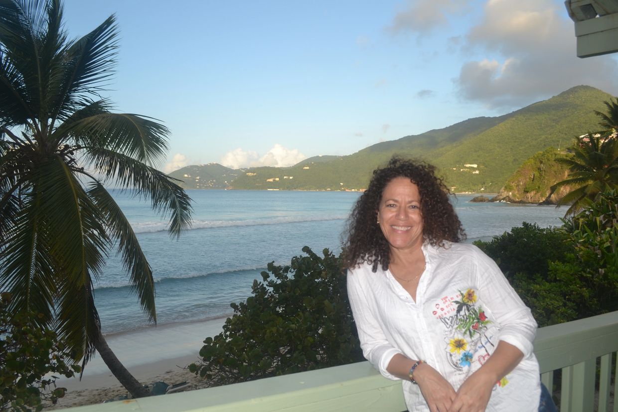 Paula smiling by the Tortola North Shore seaside with palm trees and mountains in the background.