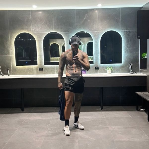 A tattooed man taking a gym mirror selfie with headphones on.