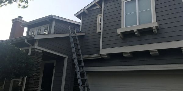 2 story gray paneled home with a ladder balanced on the roofline