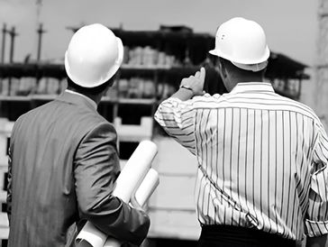 Two men in hard hats analyze a construction site, one gesturing and the other holding blueprints
