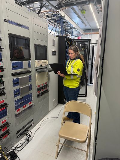 Woman in yellow safety jacket working on laptop in server room.