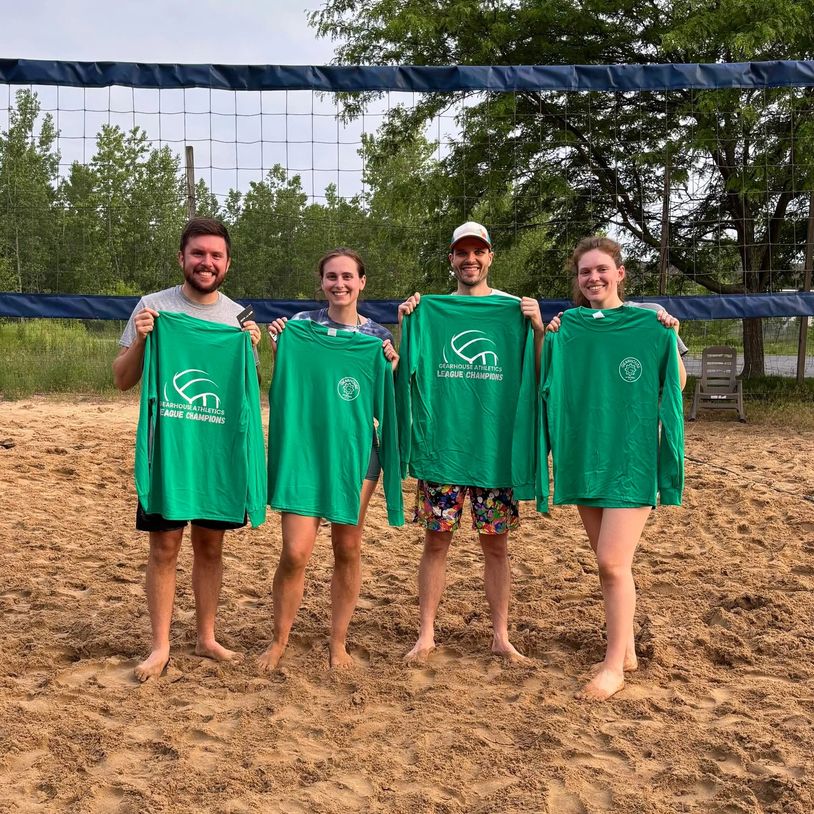 Four people proudly hold green championship shirts on a sandy volleyball court.