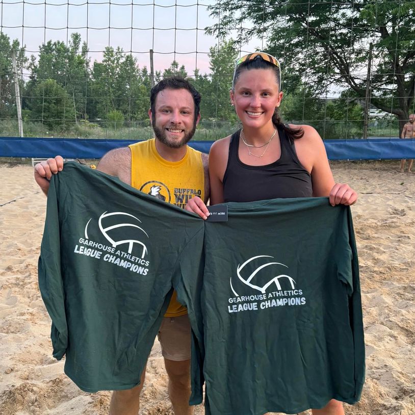 Two volleyball players proudly hold "League Champions" shirts on a sandy court.