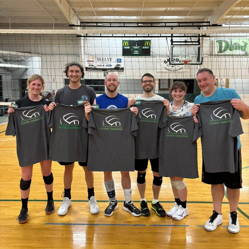 Six volleyball players proudly hold championship t-shirts on a gym court.