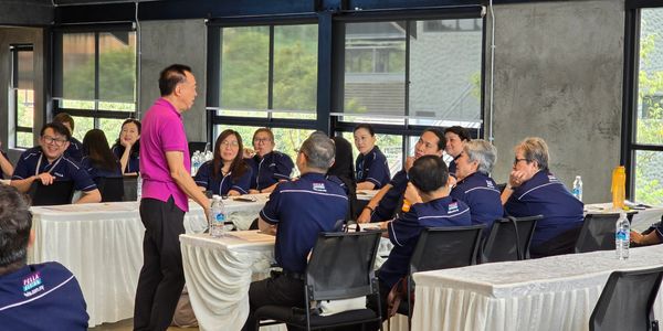 A man in a pink shirt speaking to a group seated in navy uniforms during a workshop.