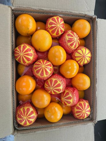 Box of fresh oranges, some wrapped in red and yellow paper.