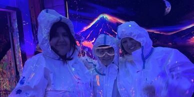 Three people in white suits under UV light splattered with neon paint.