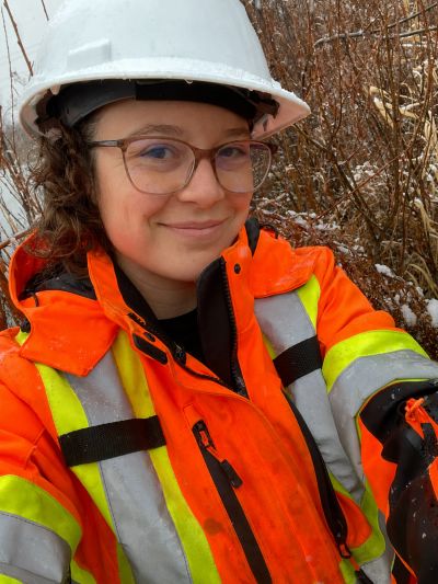 Woman in safety gear smiling outdoors in a snowy, wooded area.