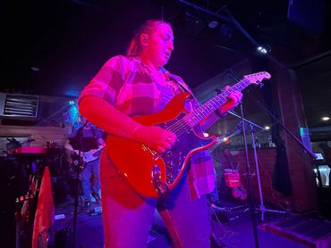 Woman playing electric guitar on stage under purple lighting.