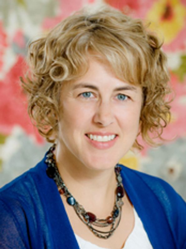 Woman with curly blonde hair wearing a blue top and necklace, smiling.