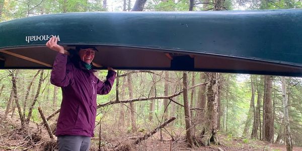 Person carrying a canoe in a forested area, smiling.