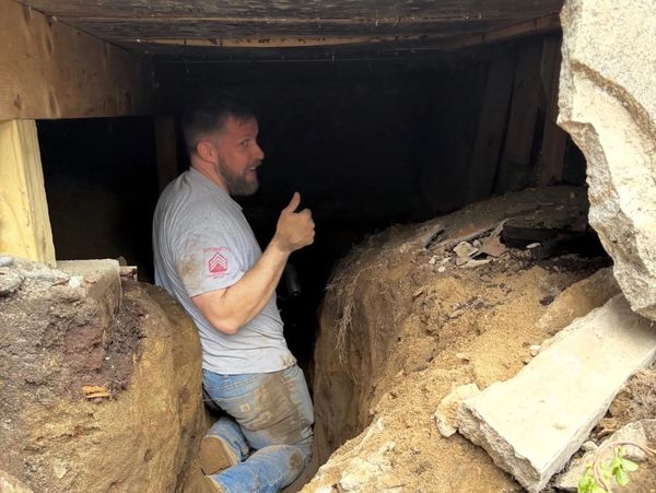 Man giving thumbs up while kneeling in a muddy trench inside a wooden structure.