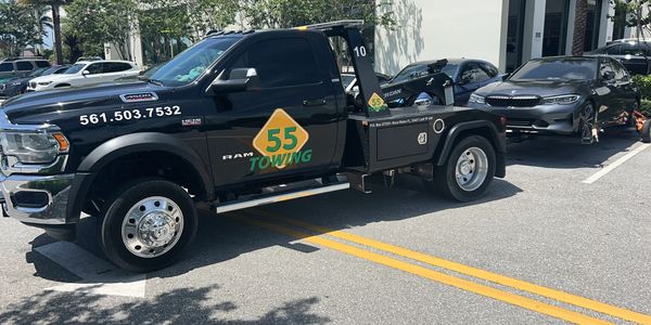 55 Towing is hauling a gray BMW sedan in a parking lot. The truck is positioned in front of the car, with the BMW secured on its lift. The background shows other parked vehicles and typical parking lot surroundings.