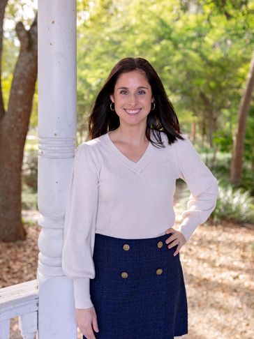 Smiling woman stands outdoors next to a white pillar with greenery in the background.