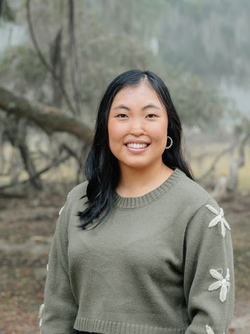 Smiling woman in a green sweater outdoors with blurred trees in the background.