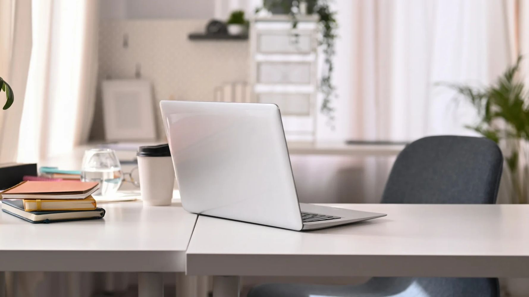 A sleek laptop on a bright desk with notebooks and a coffee cup nearby.