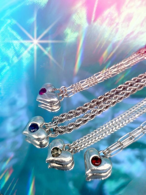 Four silver heart necklaces with colorful gems on shimmering blue background.