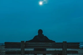 A person sitting alone on a bench under a bright full moon at night.