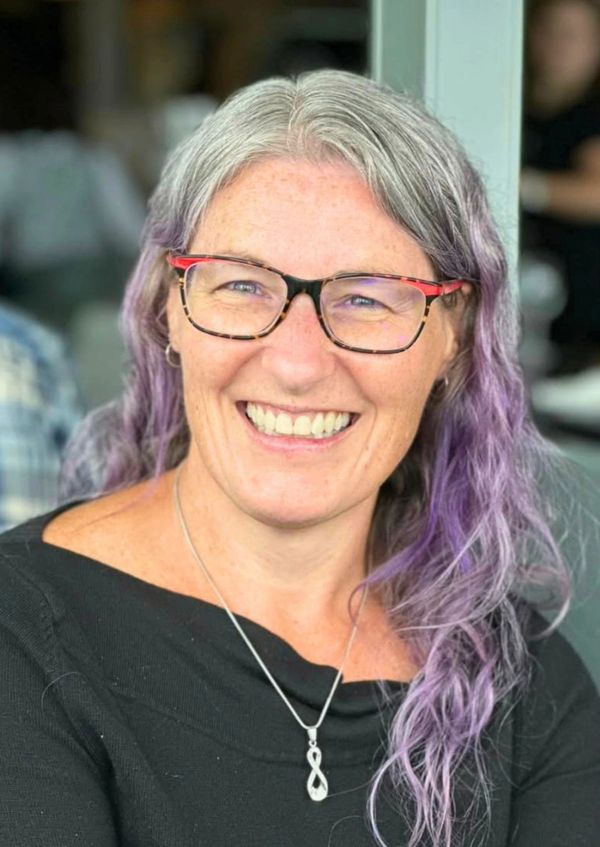 Smiling woman with gray and purple hair wearing glasses and a black top.