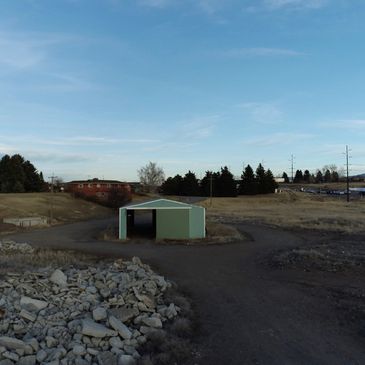 Small green shed in a rural landscape with rocky ground and trees.