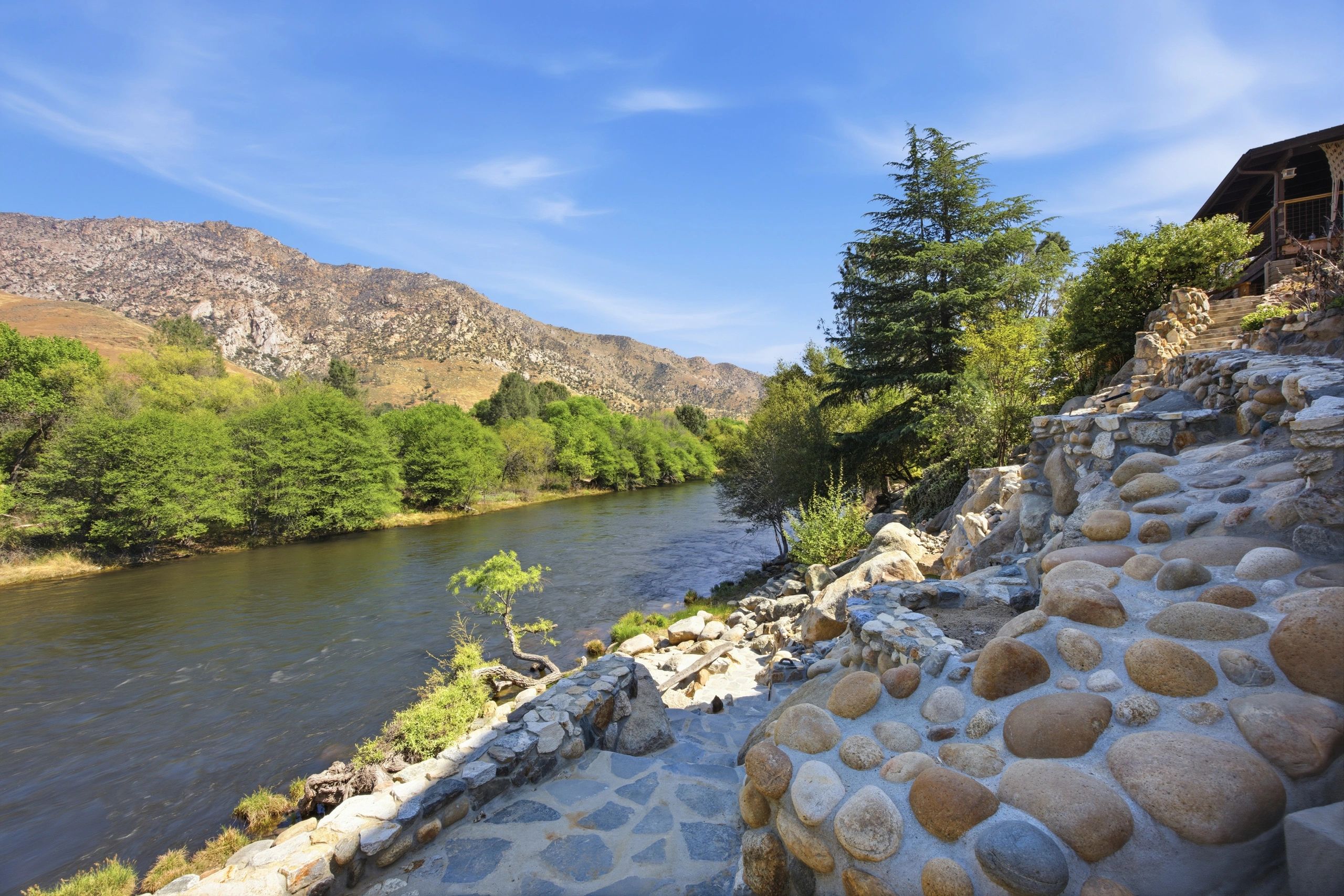 Stone pathway beside a river with mountains and trees under a clear blue sky.