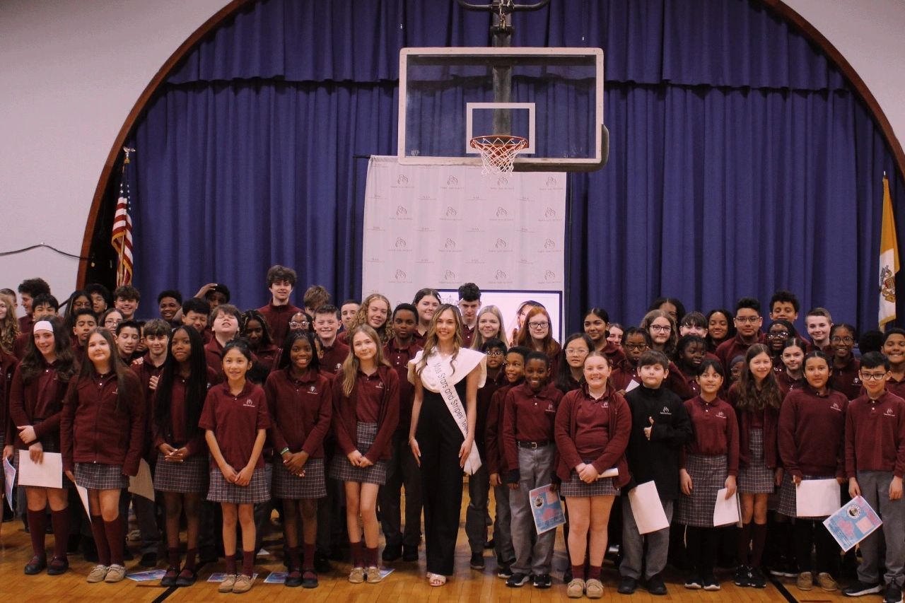 Group photo of students and a woman wearing a sash in a school gym.
