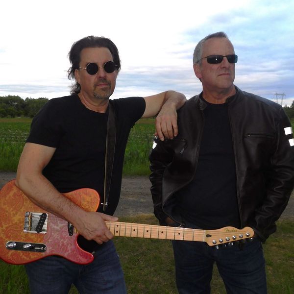 Two men with sunglasses, one holding an electric guitar, standing outdoors.