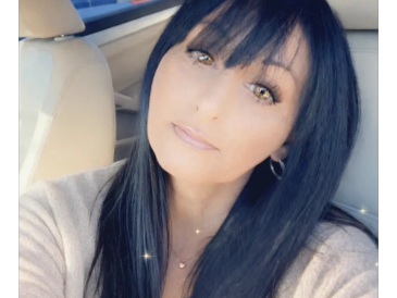 Woman with long black hair sitting in a car, looking at the camera.