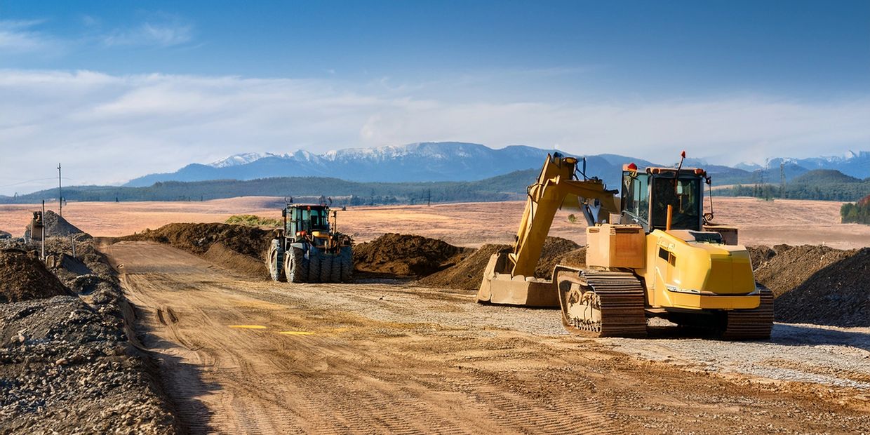 Construction vehicles working on a dirt road with mountains in the background.
