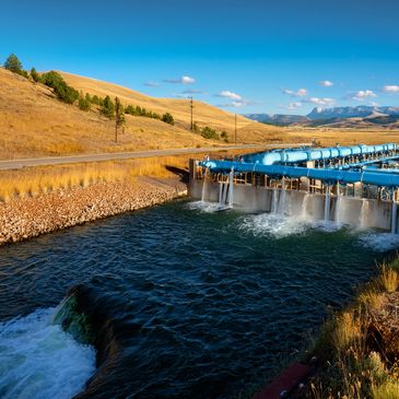 Water flowing through a dam in a rural landscape with hills and a clear sky.
