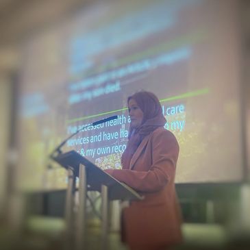 A woman in a hijab delivering a speech at a podium with a blurred presentation behind her.