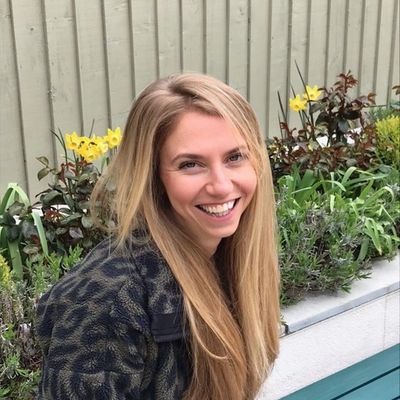 A smiling woman with long blonde hair outdoors near flowers.