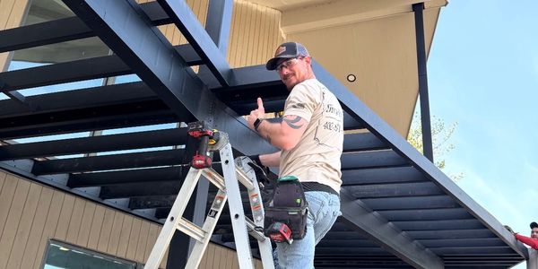 Man giving a thumbs-up on a ladder while working outdoors.