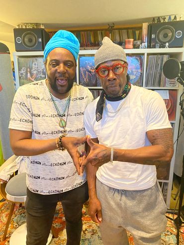 Two men posing together in a music studio, one in a blue headwrap and the other in a gray beanie.