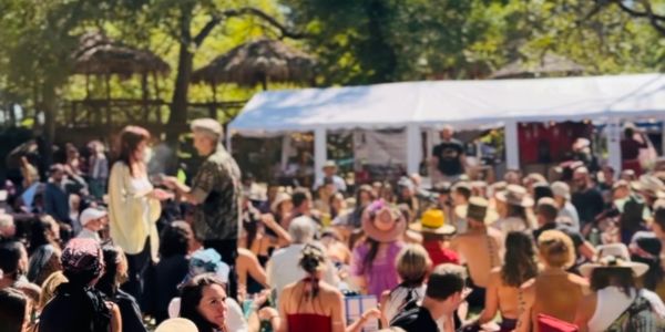 Crowd gathered outdoors at a sunny festival with tents and trees.