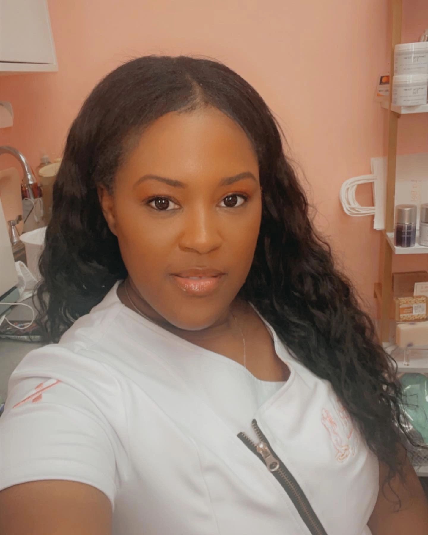 Confident woman in a white medical uniform taking a selfie in a peach-toned room.