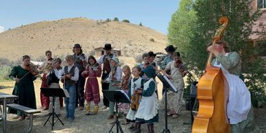 Group of people in pioneer clothing playing string instruments outdoors.