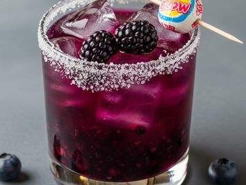 A vibrant purple cocktail with blackberries, ice, and a Blow Pop garnish on a salted rim glass.
