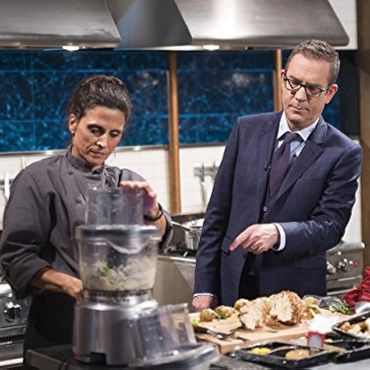 A chef uses a food processor while a man in a suit watches.
