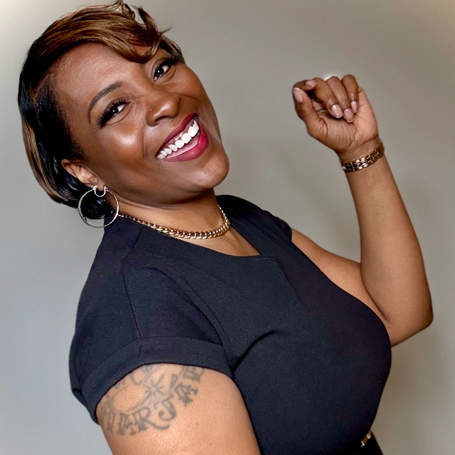 Smiling woman in a black dress with a tattoo and gold jewelry.