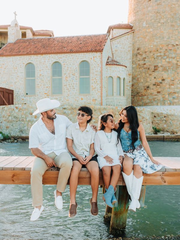 Family of four sitting on dock near stone building and water.