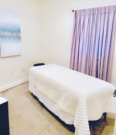 A serene massage room with a white quilted massage table and lavender curtains.