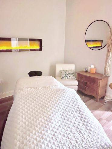 Cozy massage room with a white treatment table, chair, and warm fireplace ambiance.