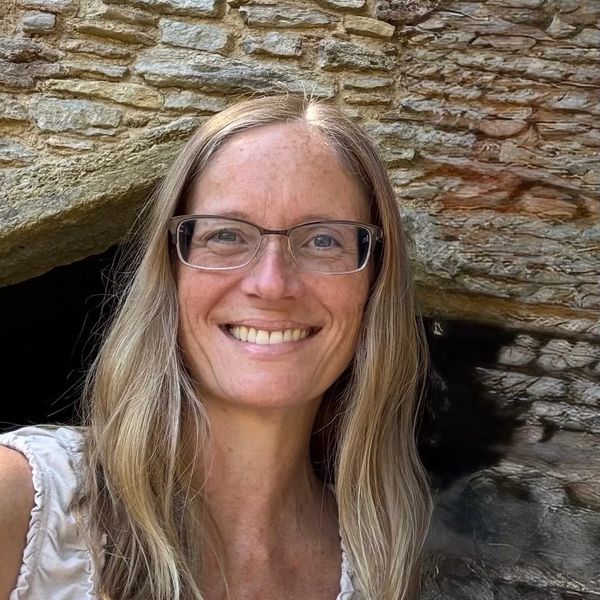 Smiling woman with glasses in front of a textured stone wall.