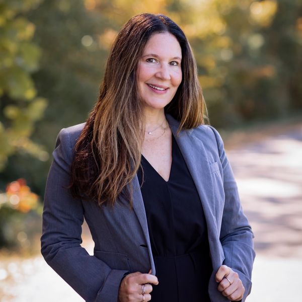Confident woman in business attire smiles outdoors with blurred greenery background.